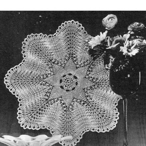 May include: A white crocheted doily with a delicate lace pattern. The doily is displayed on a table with a glass vase of flowers and a clear glass bowl.