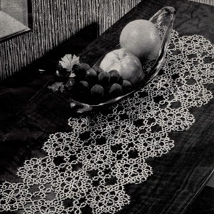 Op de afbeelding: Een witte gehaakte tafelloper met een delicaat bloemenpatroon. De loper ligt op een donkerhouten tafel met een glazen kom met fruit erop.