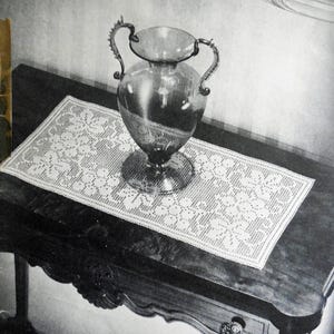 May include: A white crocheted table runner with a delicate floral pattern sits on a dark wooden table with a carved edge. A clear glass vase with ornate handles sits on the runner.