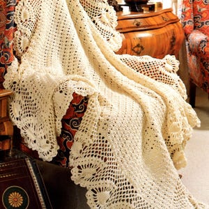 May include: A crocheted throw blanket in a cream color with a delicate floral pattern. The blanket is draped over a chair with a floral patterned fabric.