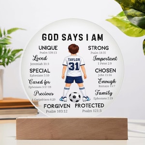 May include: A circular acrylic plaque on a rectangular wooden base featuring a cartoon soccer player wearing a white jersey with the name Taylor and number 31. The text reads God Says I Am followed by religious affirmations and scripture citations.