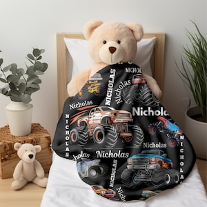 May include: A black blanket featuring colorful monster truck designs and the name "Nicholas" in white. The blanket is draped over a teddy bear on a bed. The trucks are in shades of orange, blue, and yellow.
