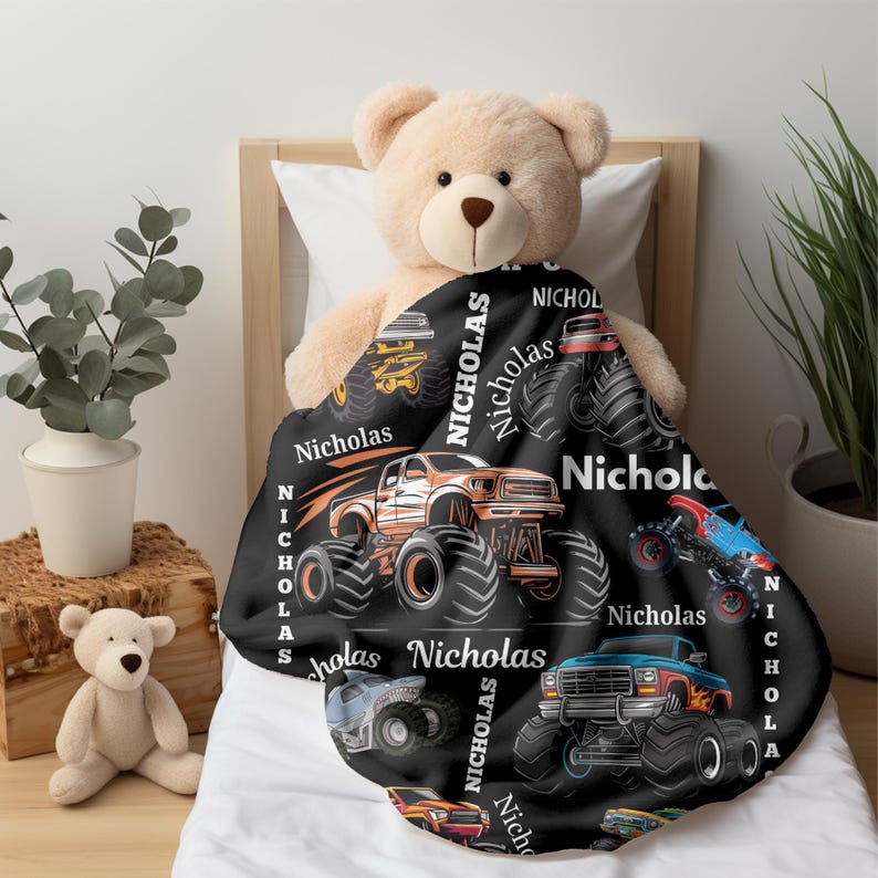 May include: A black blanket featuring colorful monster truck designs and the name "Nicholas" in white. The blanket is draped over a teddy bear on a bed. The trucks are in shades of orange, blue, and yellow.