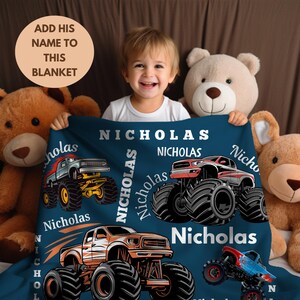 May include: A navy blue blanket featuring monster truck graphics and the name "Nicholas" in white. The blanket has illustrations of various monster trucks in red, orange, blue, and gray. The text "Add his name to this blanket" is also visible.