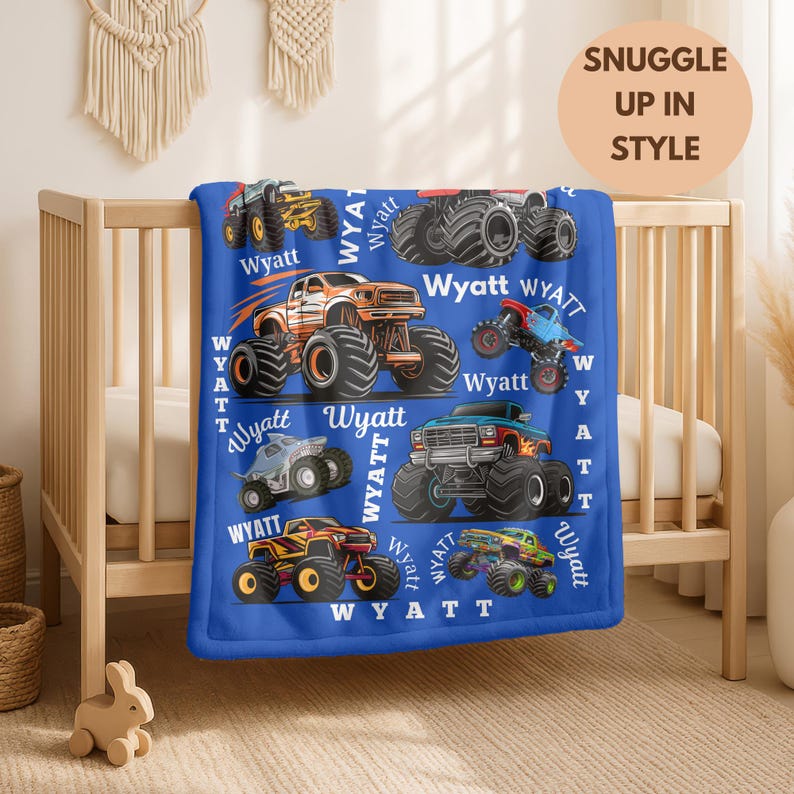 May include: A blue blanket featuring colorful monster truck illustrations and the name "Wyatt" repeated throughout. The blanket has a soft texture and is displayed on a wooden crib. The text "SNUGGLE UP IN STYLE" is also visible.