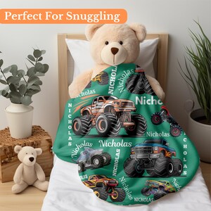 May include: A green blanket featuring illustrations of monster trucks in various colors, including orange, blue, and yellow. The blanket has the name "Nicholas" printed on it. The text "Perfect For Snuggling" is at the top.