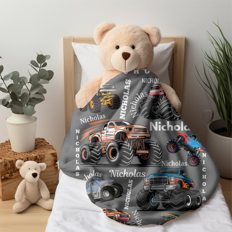 May include: A gray blanket featuring colorful monster truck illustrations and the name "Nicholas" in white. The blanket showcases various monster truck designs in orange, blue, yellow, and red, with large tires.
