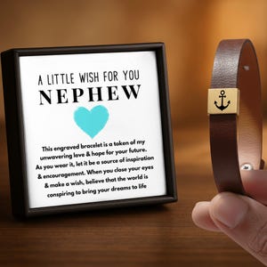 Könnte beinhalten: Ein braunes Lederarmband mit einem quadratischen goldfarbenen Metallanhänger mit eingraviertem schwarzem Ankersymbol, präsentiert neben einer quadratischen Geschenkbox mit dem Text A LITTLE WISH FOR YOU NEPHEW und einem hellblauen Herz.