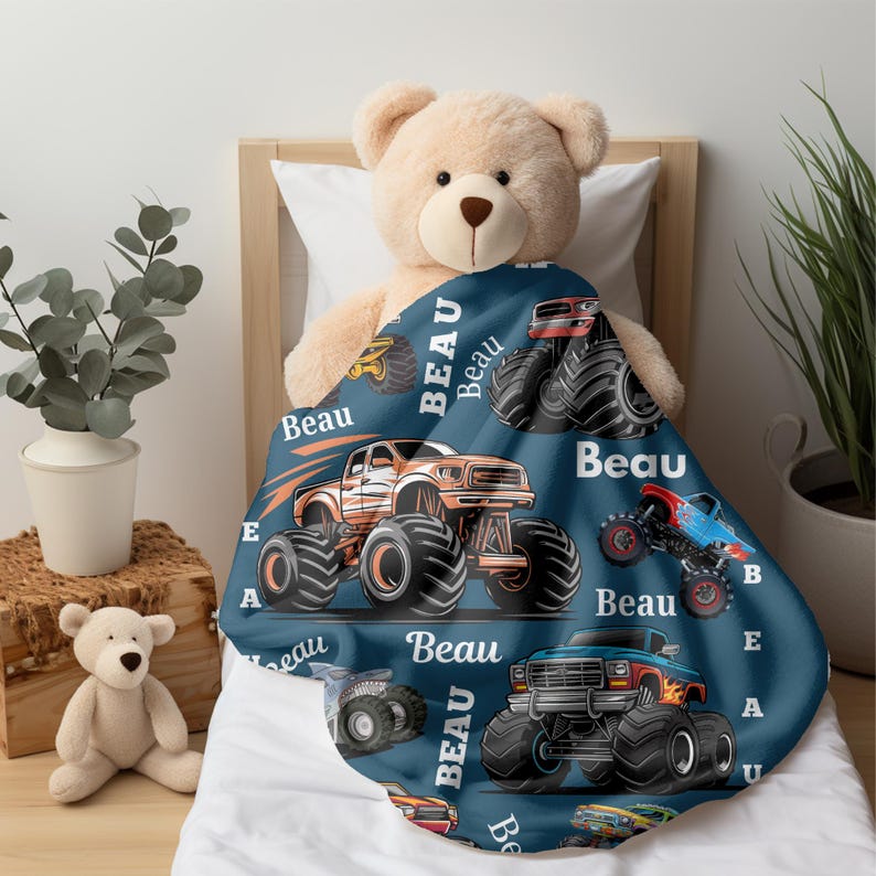 May include: A navy blue blanket featuring illustrations of monster trucks in orange, red, and blue. The word "Beau" is printed in white across the blanket. The blanket is draped over a teddy bear on a bed.