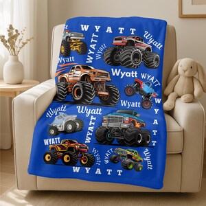 May include: A blue blanket featuring colorful monster truck illustrations. The blanket has the name "Wyatt" printed in white script throughout the design. The trucks are in various colors, including red, orange, and blue.