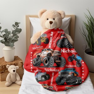 May include: A red blanket featuring monster truck illustrations in various colors, with the name "Nicholas" printed on it. The blanket is draped over a bed with a teddy bear.