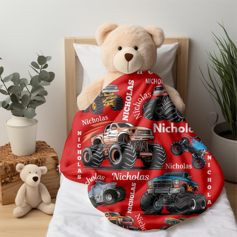 May include: A red blanket featuring monster truck illustrations in various colors, with the name "Nicholas" printed on it. The blanket is draped over a bed with a teddy bear.