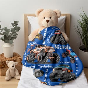 May include: A blue blanket featuring colorful monster truck illustrations and the name "Nicholas" in white. The blanket is draped over a teddy bear on a bed. The trucks are in various colors, including orange, red, and blue.
