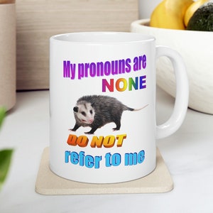 Peut inclure: Une tasse en céramique blanche avec un graphique d'un opossum et le texte "Mes pronoms sont AUCUN Ne me référez pas". Le texte est en couleurs arc-en-ciel.