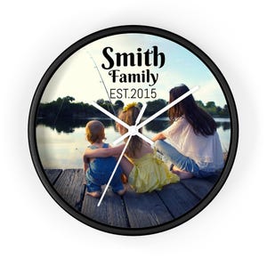 May include: A black and white photo of a family of three sitting on a wooden dock by a lake. The photo is on a round clock with black hands and a black frame. The text "Smith Family EST. 2015" is printed on the clock.