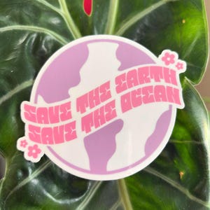 May include: A circular sticker featuring a light purple globe design with the pink text "SAVE THE EARTH SAVE THE OCEAN". The sticker is decorated with small flower accents and is set against a dark green leaf.