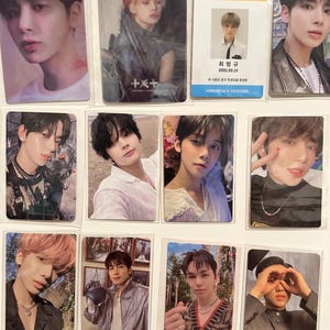 May include: A collection of 12 photo cards featuring a K-pop boy band. The cards are printed with various images of the band members, including close-ups of their faces and full-body shots. The cards are all rectangular in shape and have a white border. The cards are arranged in a grid pattern on a white surface. The text on the cards reads "TXT" and "TOMORROW X TOGETHER".
