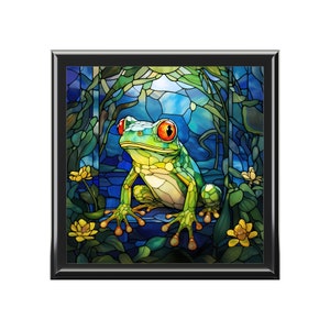 May include: A stained glass artwork depicting a green tree frog with red eyes sitting on a blue rock surrounded by green leaves and yellow flowers.