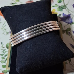 Peut inclure: Un ensemble de quatre bracelets joncs en métal argenté poli à la surface lisse et arrondie. Les bracelets sont empilés ensemble et présentés sur un coussin rectangulaire en velours noir, mettant en valeur une finition métallique réfléchissante.