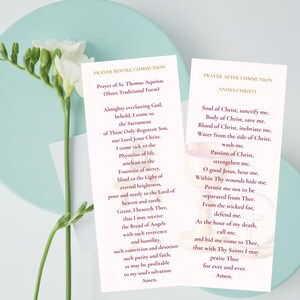 Puede incluir: Dos tarjetas de oración con texto religioso, "Oración antes de la comunión" y "Oración después de la comunión", se muestran sobre una superficie azul claro. Las tarjetas son blancas con texto dorado. Una ramita de flores blancas y hojas verdes está en el fondo.