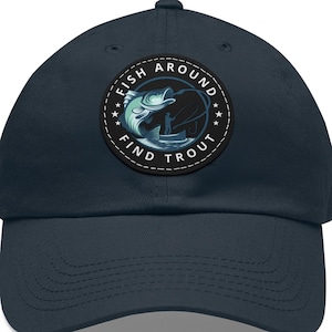以下が含まれることがあります： 紺色の野球帽。円形のパッチには、水から飛び出すマスが描かれています。パッチには「Fish Around Find Trout」の文字が円形にデザインされています。帽子は柔らかい素材でできており、つばがカーブしています。