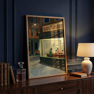 May include: A framed print of a painting depicting a diner scene with the text "Bob's Burgers" above the entrance. The artwork features a warm color palette and a street scene with figures inside the diner.