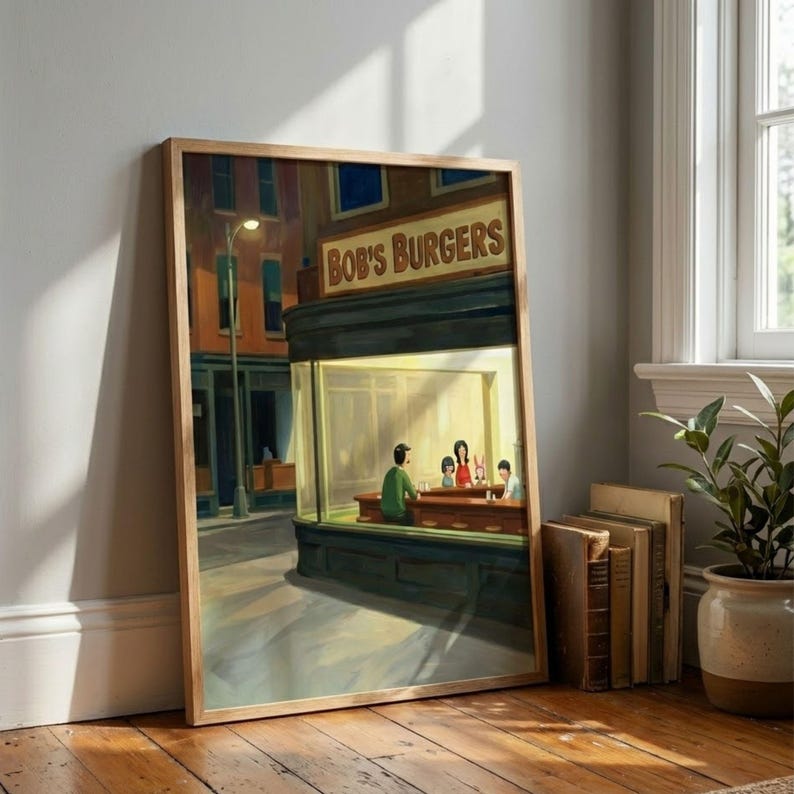 May include: A framed art print of a diner scene, with the text "Bob's Burgers" above the window. The artwork shows a nighttime street scene with figures inside the diner, lit by warm light. The frame is a light wood colour.