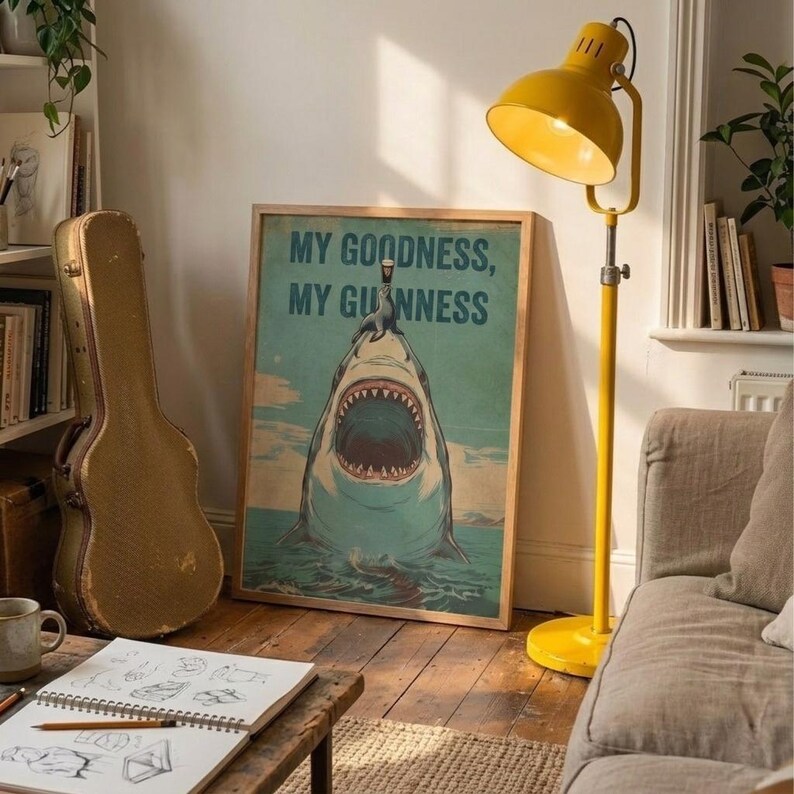 May include: A framed poster with the text "MY GOODNESS, MY GUINNESS" above an illustration of a shark with an open mouth. A yellow floor lamp and a guitar case are also visible in the room.