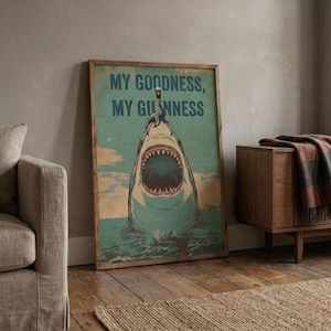 May include: A framed vintage-style poster featuring a shark with an open mouth, a seal balancing a pint of Guinness on its nose, and the text "MY GOODNESS, MY GUINNESS" in a teal background. The poster is in a wooden frame.