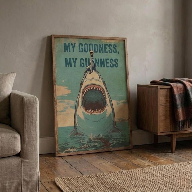 May include: A framed vintage-style poster featuring a shark with an open mouth, a seal balancing a pint of Guinness on its nose, and the text "MY GOODNESS, MY GUINNESS" in a teal background. The poster is in a wooden frame.
