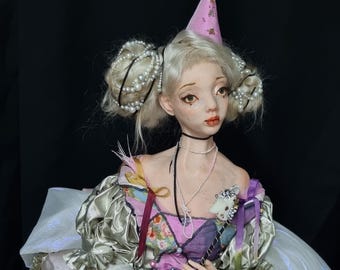 Puede incluir: Una muñeca de porcelana que lleva un vestido multicolor con un gorro de fiesta rosa y blanco. La muñeca tiene el pelo rubio recogido en dos moños con accesorios de pelo de perlas. La muñeca sostiene una varita con una cabeza de gato blanca en la parte superior.