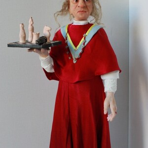 May include: A doll wearing a red dress and a blue and yellow sash, holding a small chessboard with chess pieces.