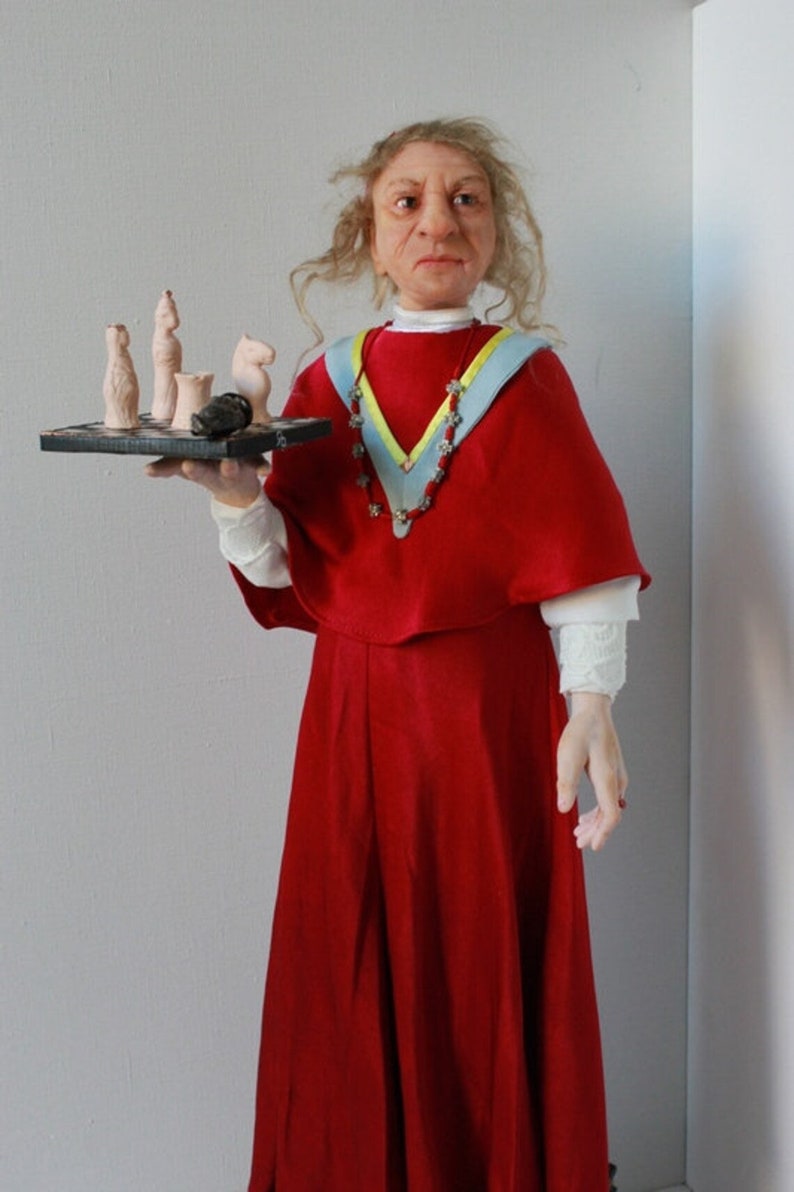 May include: A doll wearing a red dress and a blue and yellow sash, holding a small chessboard with chess pieces.
