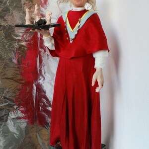 May include: A doll wearing a red robe with a blue and yellow collar and a necklace of stars. The doll is holding a tray with chess pieces on it. The doll is standing on a stack of books.