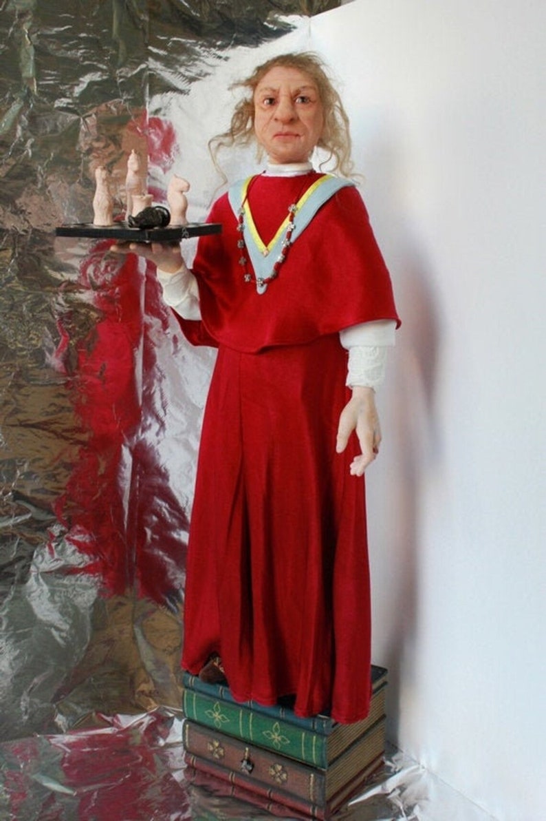 May include: A doll wearing a red robe with a blue and yellow collar and a necklace of stars. The doll is holding a tray with chess pieces on it. The doll is standing on a stack of books.