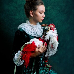 Pode incluir: Uma mulher com um vestido de veludo verde com um colar de renda branca segura uma figura de galo vermelha e dourada. O galo est&aacute; vestido com um colar de renda branca e um tecido vermelho.