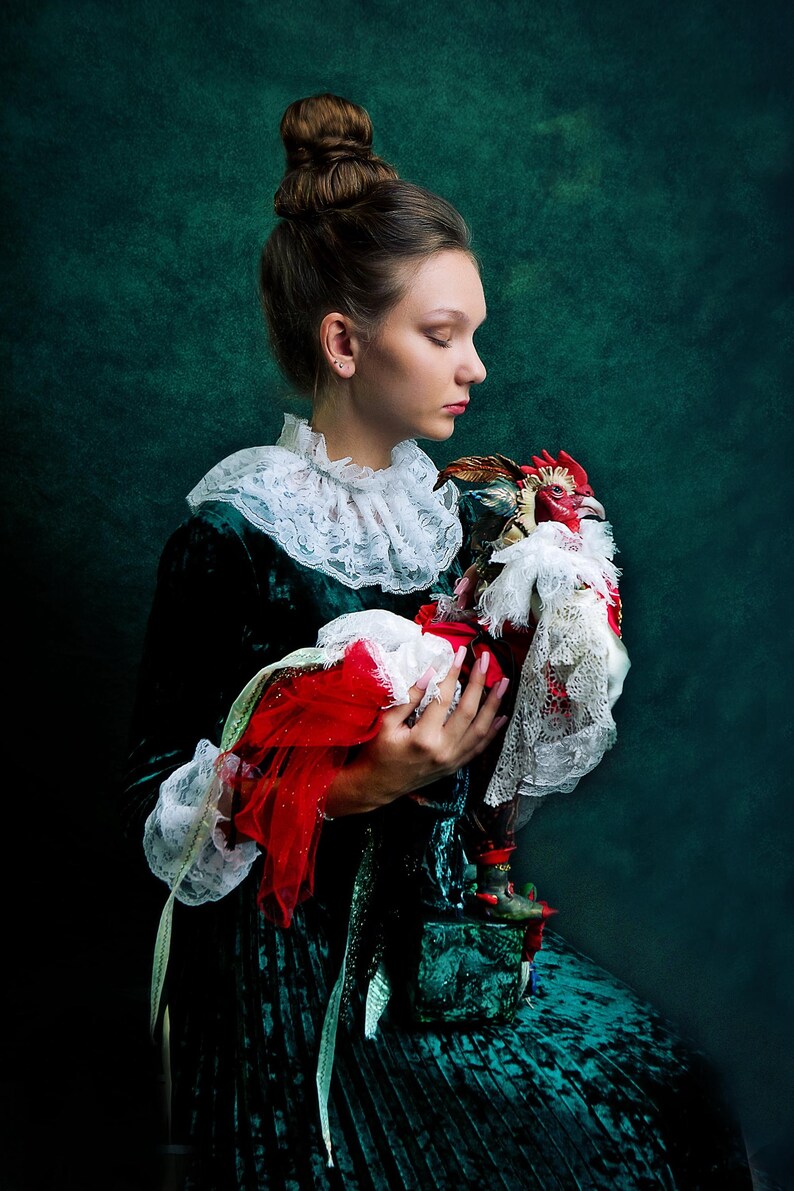Pode incluir: Uma mulher com um vestido de veludo verde com um colar de renda branca segura uma figura de galo vermelha e dourada. O galo est&aacute; vestido com um colar de renda branca e um tecido vermelho.