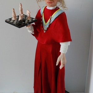 May include: A doll wearing a red robe with a blue and yellow sash and a beaded necklace. The doll is holding a small chessboard with chess pieces on it. The doll is standing on a stack of books.