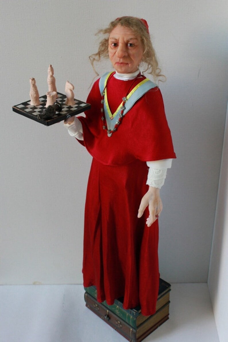 May include: A doll wearing a red robe with a blue and yellow sash and a beaded necklace. The doll is holding a small chessboard with chess pieces on it. The doll is standing on a stack of books.