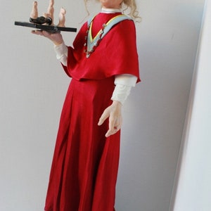 May include: A doll wearing a red dress and a white lace collar, holding a tray with small figures. The doll is standing on a stack of books.