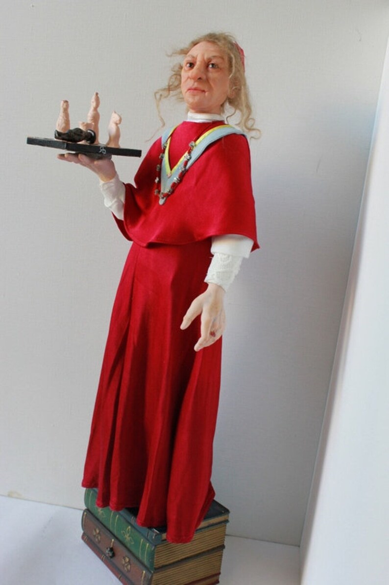 May include: A doll wearing a red dress and a white lace collar, holding a tray with small figures. The doll is standing on a stack of books.