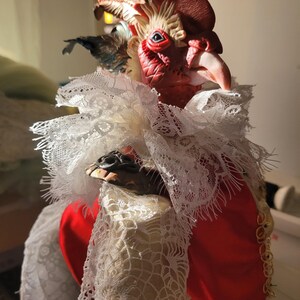 Pode incluir: Uma figura de galo vermelho com um colar de renda branca e uma corrente dourada. O galo tem uma cabe&ccedil;a detalhada com uma crista e uma barbela vermelhas, e seus olhos s&atilde;o pintados de azul.