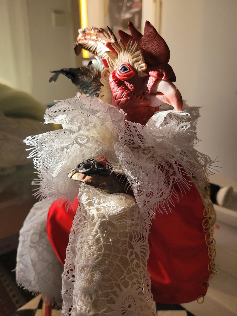 Pode incluir: Uma figura de galo vermelho com um colar de renda branca e uma corrente dourada. O galo tem uma cabe&ccedil;a detalhada com uma crista e uma barbela vermelhas, e seus olhos s&atilde;o pintados de azul.