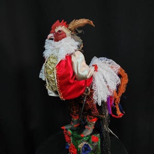 Pode incluir: Uma figura de galo vermelho, branco e dourado vestindo uma jaqueta vermelha, um colar de renda branca e cal&ccedil;as pretas e vermelhas. O galo est&aacute; em p&eacute; sobre uma base verde, vermelha e azul com flores.