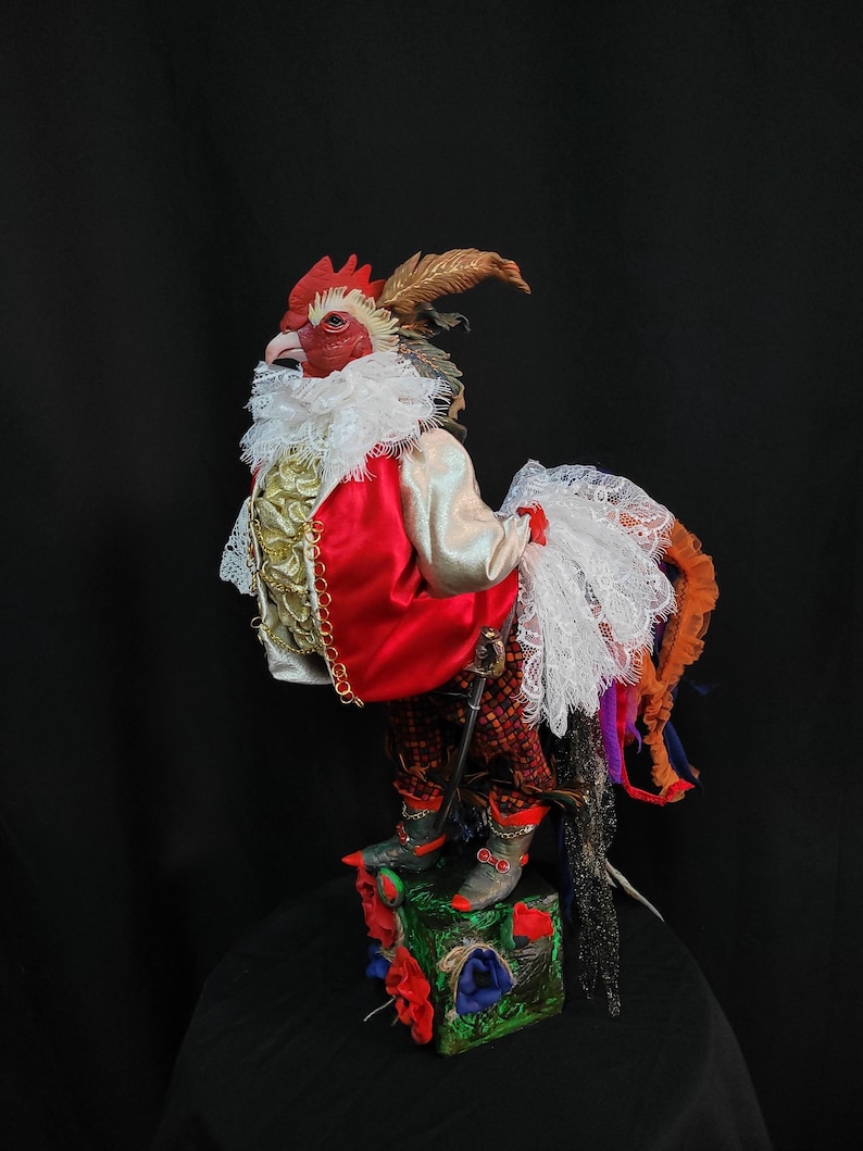Pode incluir: Uma figura de galo vermelho, branco e dourado vestindo uma jaqueta vermelha, um colar de renda branca e cal&ccedil;as pretas e vermelhas. O galo est&aacute; em p&eacute; sobre uma base verde, vermelha e azul com flores.