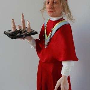 May include: A doll dressed as a cardinal holding a small chessboard with a black and white chequered pattern. The doll has long curly hair and is wearing a red robe with a blue and yellow sash. The chessboard has a few chess pieces on it, including a black pawn and a few white pawns.
