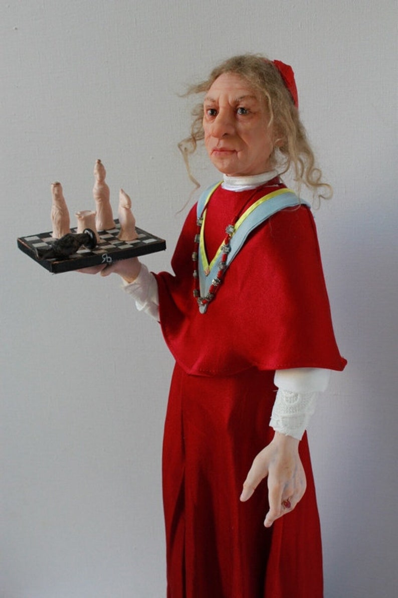 May include: A doll dressed as a cardinal holding a small chessboard with a black and white chequered pattern. The doll has long curly hair and is wearing a red robe with a blue and yellow sash. The chessboard has a few chess pieces on it, including a black pawn and a few white pawns.