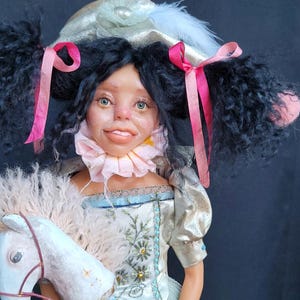 May include: A handcrafted doll with black pigtails, dressed in a silver and blue gown, clutching a white toy horse. The doll features a pink frilled collar and a hat adorned with feathers and pink ribbons. The doll's face has a distinctive design.