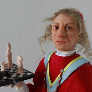 May include: A doll wearing a red robe with a blue and yellow sash, holding a chessboard with white chess pieces and a black chess piece.