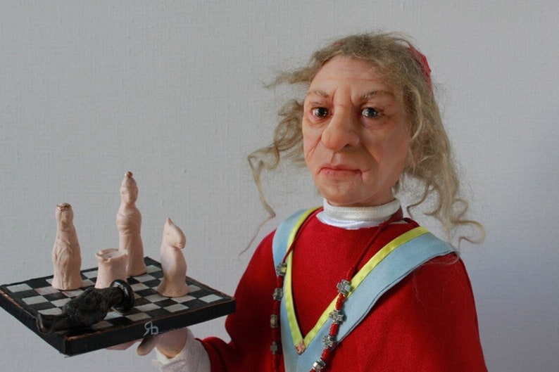 May include: A doll wearing a red robe with a blue and yellow sash, holding a chessboard with white chess pieces and a black chess piece.